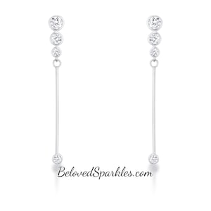 May include: A pair of silver earrings with a linear design. Each earring features three graduated round, clear gemstones connected to a long, straight silver bar, with a single gemstone at the end. The website BelovedSparkles.com is printed at the bottom.
