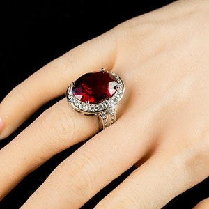 Può includere: Un anello d'argento con una grande pietra rossa ovale circondata da pietre chiare più piccole.