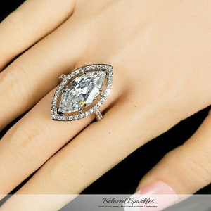 Op de afbeelding: Een ring met een marquise-geslepen diamant met een halo van kleinere diamanten. De ring is gezet op een zilveren band en wordt getoond op een hand met een zwarte achtergrond. De ring is een sieraad.