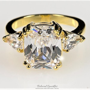 Puede incluir: Un anillo de oro con una gran piedra de circonita cúbica cuadrada, transparente, rodeada de dos piedras de circonita cúbica más pequeñas en forma de pera.