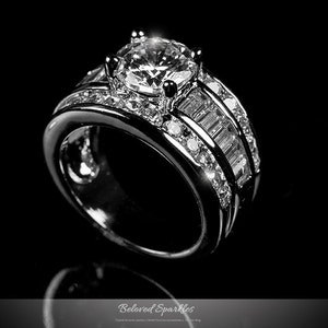 Puede incluir: Un anillo de plata con un gran diamante redondo y claro en el centro y una banda de diamantes más pequeños. La banda está engastada con diamantes de forma baguette.