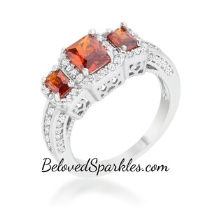 Op de afbeelding: Een zilveren ring met drie oranje edelstenen gezet in een halo van kleinere heldere stenen. De ring heeft een delicaat filigraan ontwerp.