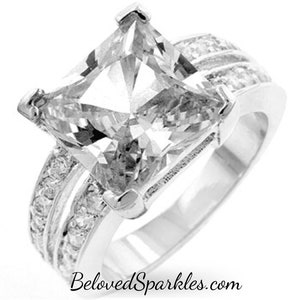 May include: A silver ring with a large, square-cut, clear cubic zirconia stone. The ring has a double band with smaller clear cubic zirconia stones.