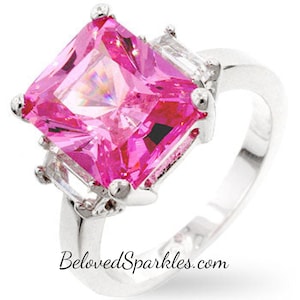 Op de afbeelding: Een zilveren ring met een grote, vierkant geslepen, helder roze edelsteen als belangrijkste kenmerk. Twee kleinere, rechthoekige, heldere edelstenen flankeren de hoofdsteen. De ringband is glad en gepolijst. De tekst "BelovedSparkles.com" staat onderaan.