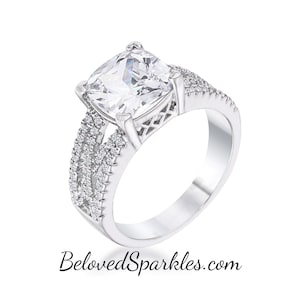 Op de afbeelding: Een zilveren ring met een grote, heldere, kussen geslepen zirkonia-steen. De ring heeft een delicaat, ingewikkeld ontwerp met kleinere zirkonia-stenen die in de band zijn gezet.