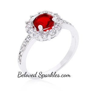 Peut inclure: Une bague en argent avec une grande pierre rouge entourée d'un halo de petites pierres blanches. La bande de la bague est également sertie de petites pierres blanches.