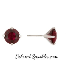 May include: A pair of stud earrings featuring a round, deep red gemstone set in a silver-colored metal. One earring is shown face-on, the other from the side, displaying the post and setting. The earrings are set against a white background.