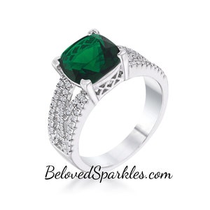 Op de afbeelding: Een zilveren ring met een grote, vierkante edelsteen in smaragdgroen. De ring heeft een delicaat, ingewikkeld ontwerp met kleinere, heldere edelstenen rond de band.