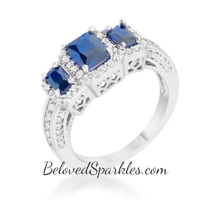 Op de afbeelding: Een zilveren ring met drie blauwe edelstenen gezet in een halo van kleinere heldere stenen. De ring heeft een delicaat filigraan ontwerp en is aan elke kant versierd met een rij heldere stenen.