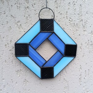 May include: A stained glass suncatcher in a diamond shape, featuring light and dark blue glass panes. The design includes black, textured squares at each corner and a metal hanging loop. The suncatcher is designed to be hung.
