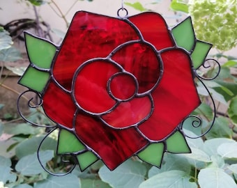 May include: A stained glass window hanging features a red rose with green leaves and black metal accents.