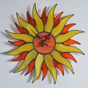 May include: A stained glass clock shaped like a sun with yellow and orange glass pieces. The clock face is a red circle with black hands.