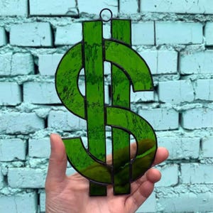 Può includere: Un simbolo del dollaro in vetro colorato verde con un contorno nero. Il simbolo del dollaro è tenuto davanti a un muro di mattoni blu chiaro. Il vetro ha un aspetto strutturato.