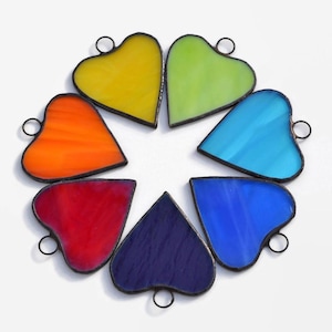 May include: Seven stained glass hearts in a rainbow color pattern. The hearts are arranged in a circle with the points of the hearts facing outward. The hearts are made of clear glass with a black outline.