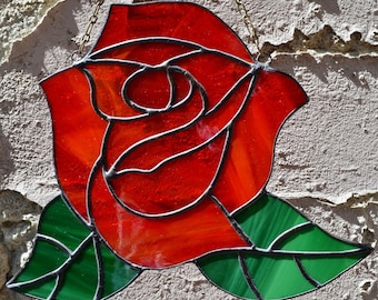 May include: A stained glass window hanging with a red rose design. The rose has a dark outline and is surrounded by two green leaves.