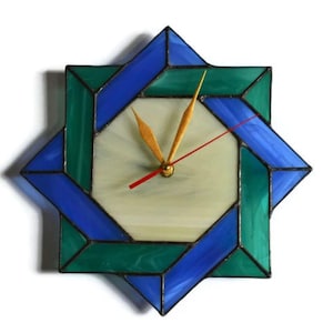 Può includere: Un orologio in vetro colorato con un design geometrico blu e verde. Il quadrante dell'orologio è bianco con lancette dorate.