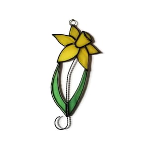 May include: A stained glass daffodil with yellow petals and green leaves. The daffodil is suspended from a wire hanger.