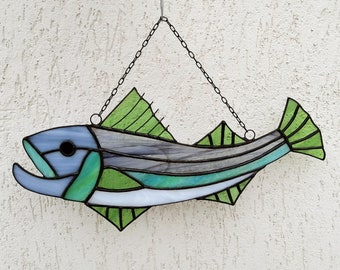 May include: A stained glass fish with green, blue, and white accents hanging from a chain. The fish has a stylized design with a long body and a pointed tail.