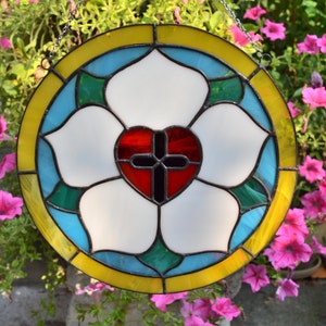 May include: A stained glass window depicting a white flower with a red heart and a black cross in the center. The flower is surrounded by a blue, green, and yellow border.