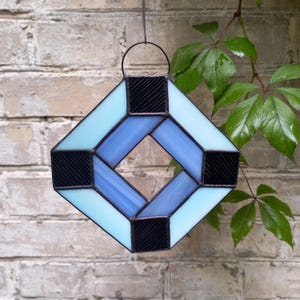 May include: A diamond-shaped stained glass suncatcher featuring a geometric pattern of light blue, medium blue, and black textured glass pieces held together by dark metal solder lines, with a circular metal loop at the top for hanging.