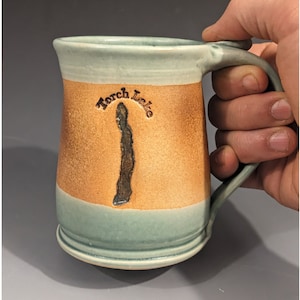 May include: Handmade ceramic mug with a light blue rim and base, and a tan middle section. The mug features the text "Torch Lake" and a dark brown outline of the lake. The handle is light blue.