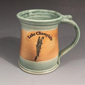 May include: A green ceramic mug with a brown band around the middle. The band has the text "Lake Champlain" and a stylized image of the lake.