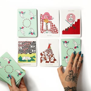 May include: A set of tarot cards with colorful illustrations. The cards feature a variety of images, including a woman in a red dress, a man with a sword, and a group of angels. The cards are laid out on a white surface.