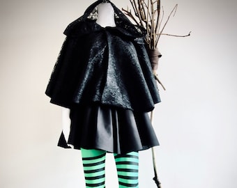 May include: A black lace cape with a black skirt and green and black striped tights. A wooden stick is leaning against a white wall.