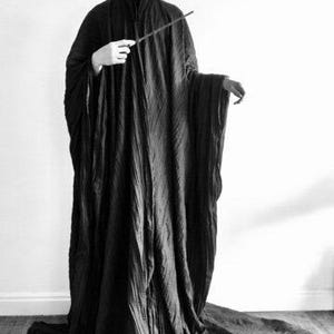 May include: A long, black robe with a flowing train, made from a textured material. The robe has long sleeves and a high collar. A wand is held in the right hand. The image is in monochrome.