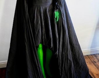 May include: A black costume with a long cape and a fitted bodice. The costume features a wide skirt and a black belt. Bright green tights and a green hand are visible. The costume is displayed on a mannequin.