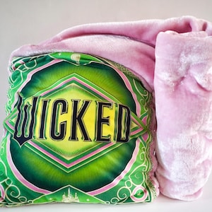 May include: A decorative pillow with the word "WICKED" in black and yellow lettering on a green background. A pink blanket is draped over the pillow. The pillow has a green and pink border with a castle design.