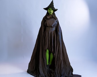 May include: A person in a dark brown witch costume with a pointed hat and long cape. The person's face is painted green, and they are wearing green tights. The costume has a high-low hemline.