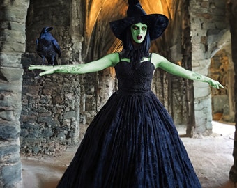 May include: A woman dressed as a witch with green face paint and a black pointed hat stands in a stone archway. She is wearing a black lace dress and holding a black raven on her outstretched arm.