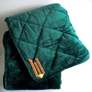 Peut inclure: Une couverture matelassée vert émeraude pliée avec un emblème brodé doré. La couverture présente un motif en losanges et est faite d'une matière douce et veloutée. L'emblème est une lettre "W" stylisée.