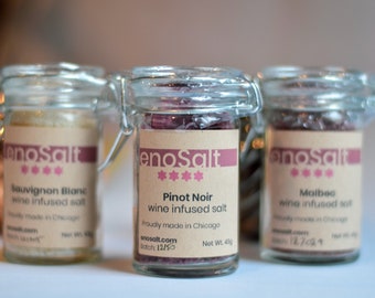 May include: Three glass jars with brown labels and silver lids containing wine infused salt. The labels read "enosalt" and list the wine varietals: Sauvignon Blanc, Pinot Noir, and Malbec. Each jar is 128 grams.