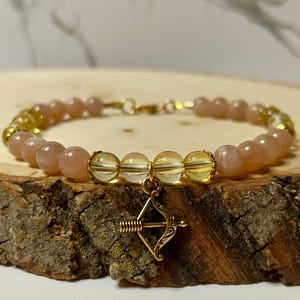 Könnte beinhalten: Ein Armband mit pfirsichfarbenen und klaren Perlen, goldfarbenen Akzenten und einem goldfarbenen Pfeil-und-Bogen-Anhänger. Das Armband wird auf einer Holzoberfläche präsentiert.