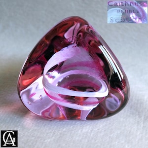 May include: A Caithness Pebble art glass paperweight. The paperweight is a triangular shape with a rounded top. It features a swirl of pink and white colours. The text "Caithness Pebble Scotland" is visible on the top right.