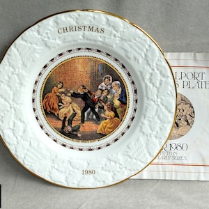 May include: A white porcelain Christmas plate from 1980 with a gold rim and embossed holly leaf border. The centre features a colourful illustration of children playing Blind Man's Buff. It is accompanied by a paper document titled Coalport Christmas Plate.