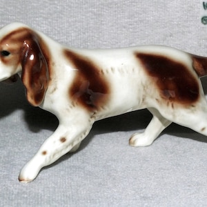 Peut inclure: Une figurine en porcelaine blanche et brune d'un chien de chasse. Le chien est en position de course, la tête tournée sur le côté. La figurine est marquée "Allemagne de l'Ouest".