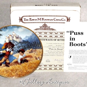 May include: A decorative plate from The Edwin M. Knowles China Co. featuring a scene from "Puss in Boots." The plate has a gold rim and depicts a cat in a hat and a boy in a field. Includes a certificate of authenticity and the text "Puss in Boots."