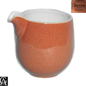 May include: A ceramic creamer jug with a speckled terracotta orange exterior and a smooth white interior. The base features a stamp with the text Denby England, dishwasher, oven safe, freezer, and microwave safe, indicating its functional use.
