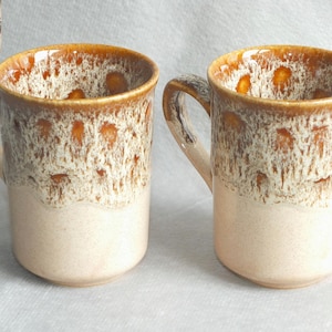 May include: Two ceramic mugs with a speckled brown and cream design. The mugs have a cylindrical shape with a handle and a light cream base. The top rim and handle are brown. The mugs are marked "Fosters Oven Dishwasher Microwave Proof".