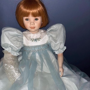 May include: A porcelain doll with short auburn hair and blue eyes, wearing a white dress with puffed sleeves, a blue sheer overlay, and a silver-toned waistband. The bodice features small blue floral embroidery. One arm is wrapped in clear bubble wrap.