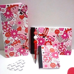 May include: A collection of notebooks and a pen. The notebooks feature a repeating pattern of smiley faces, flowers, hearts, cowboy hats, and disco balls in shades of pink, red, and white. The pen is red with a decorative top. A small handwritten note says "love".