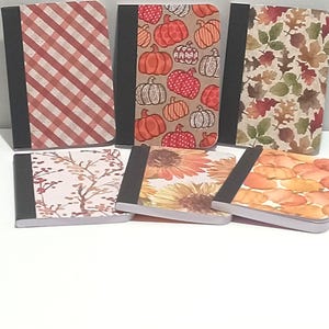 May include: A collection of small notebooks with various autumn-themed covers. Designs include pumpkins, sunflowers, fall leaves, and a red and white plaid pattern. Each notebook has a black spine.
