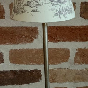 May include: A silver-toned table lamp with a cream-colored lampshade featuring a gray pastoral scene. The lamp has a slender stem and a decorative base. The background is a brick wall.