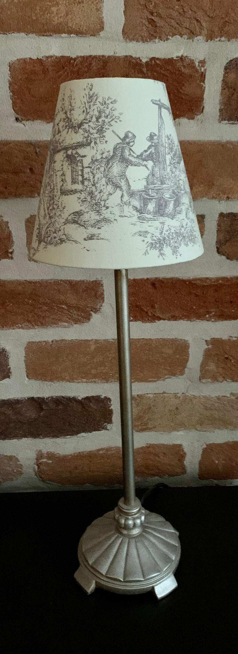 May include: A silver-toned table lamp with a cream-colored lampshade featuring a gray pastoral scene. The lamp has a slender stem and a decorative base. The background is a brick wall.