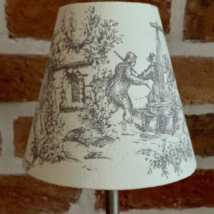 May include: Cream-colored lampshade with a black and white pastoral scene. The illustration depicts figures near a well, trees, and a building. The lampshade is cone-shaped and mounted on a silver metal stand. The background is a brick wall.