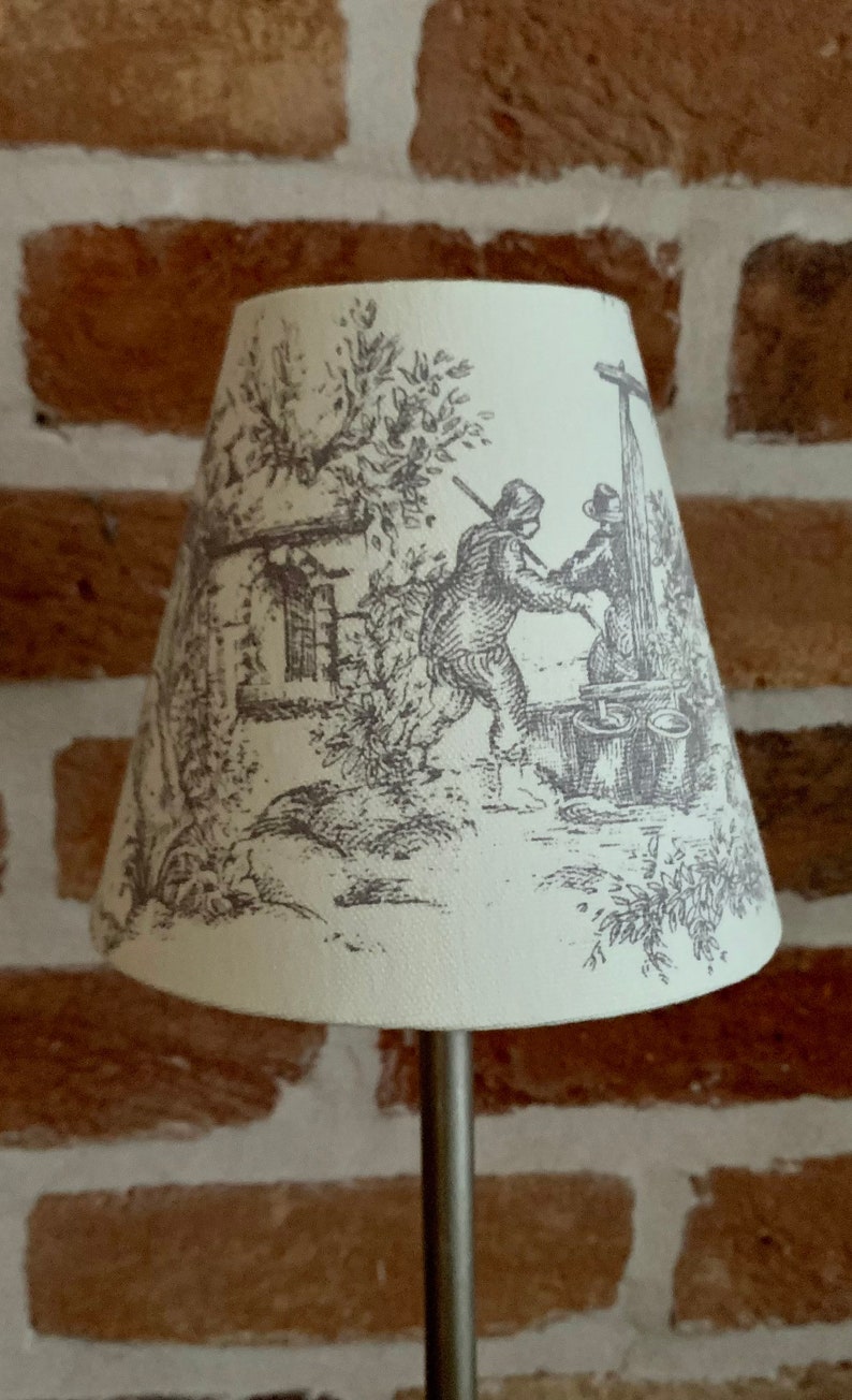 May include: Cream-colored lampshade with a black and white pastoral scene. The illustration depicts figures near a well, trees, and a building. The lampshade is cone-shaped and mounted on a silver metal stand. The background is a brick wall.