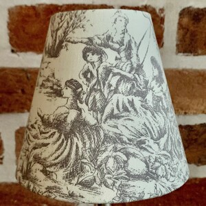 May include: A small, off-white lampshade with a detailed gray toile design featuring figures in a pastoral scene. The lampshade is cone-shaped and mounted on a silver metal stand. The background is a brick wall.